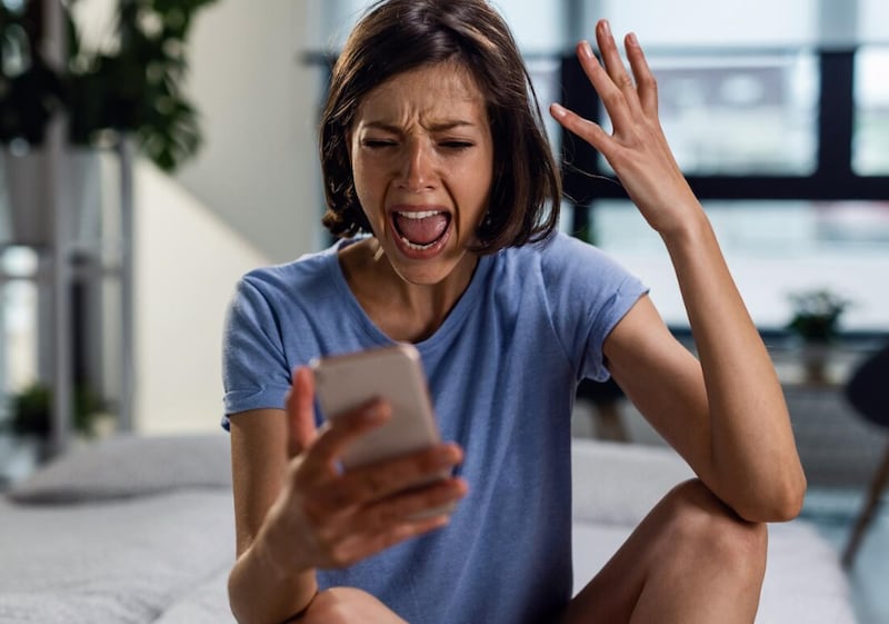 Mujer frustrada gritando a su teléfono mientras se sienta en la cama y lee el mensaje de texto que ha recibido